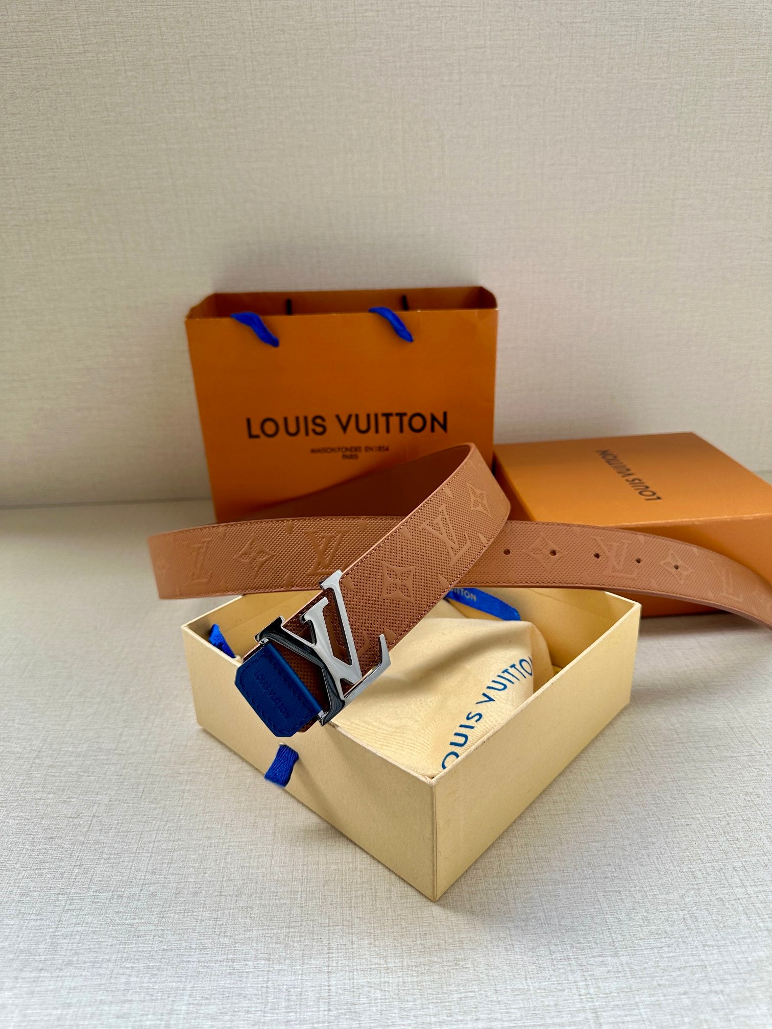 Tan monogram belt with silver LV buckle and blue keeper tab, laid out