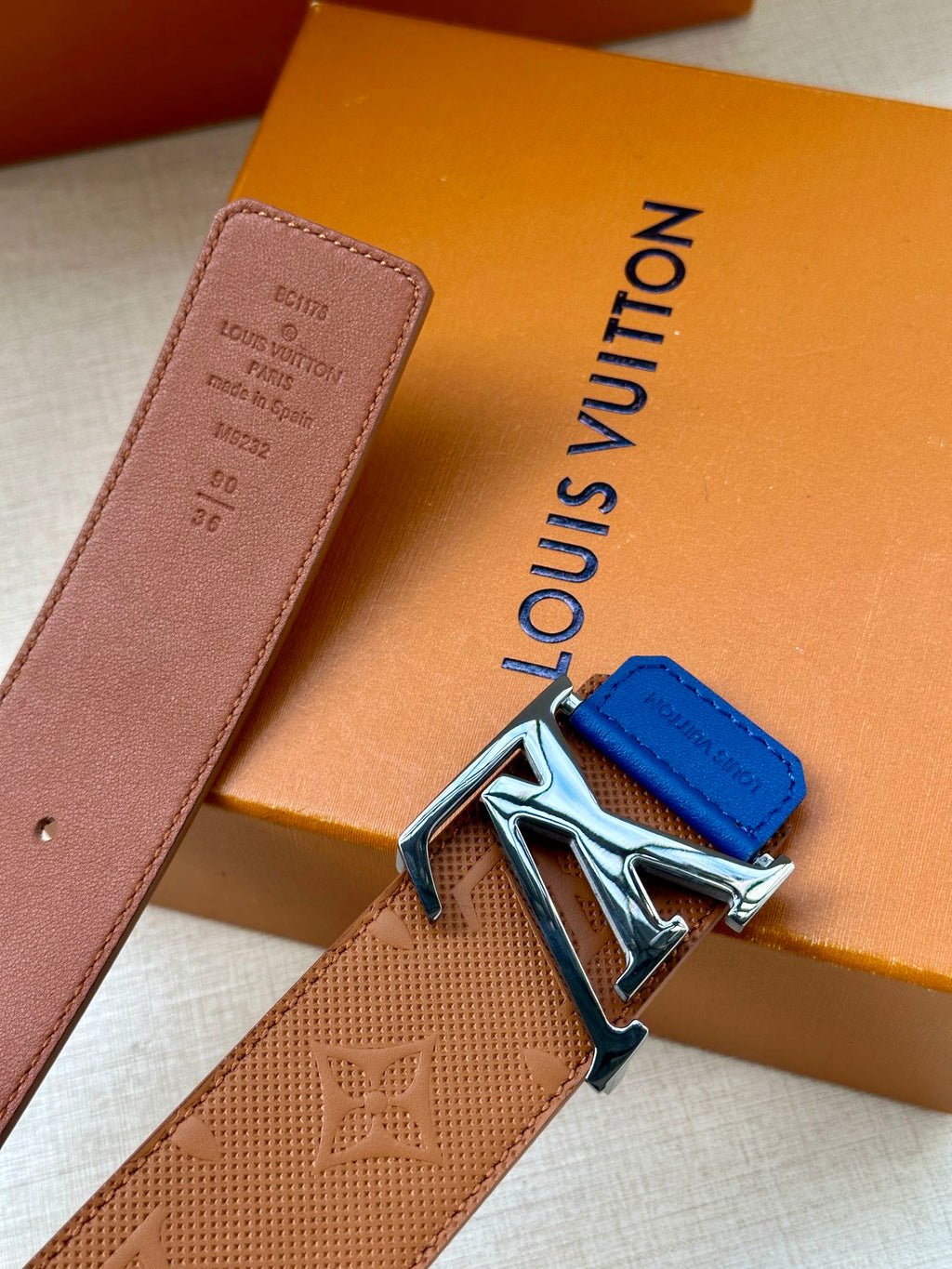 Tan monogram belt with silver LV buckle and blue keeper tab, angled view