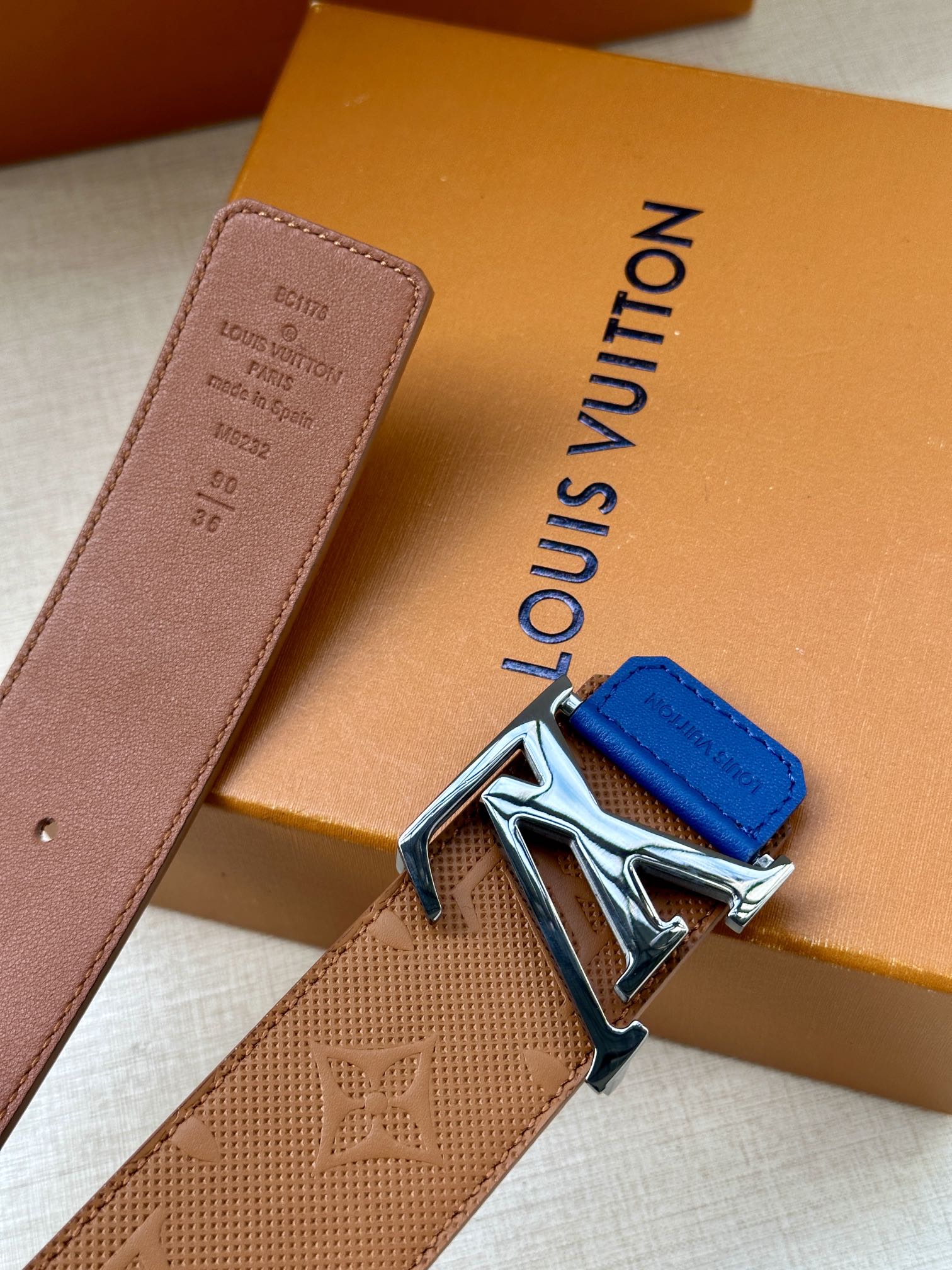 Tan monogram belt with silver LV buckle and blue keeper tab, angled view