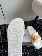 White slide sandals with textured outsole and orange logo strap on upper.