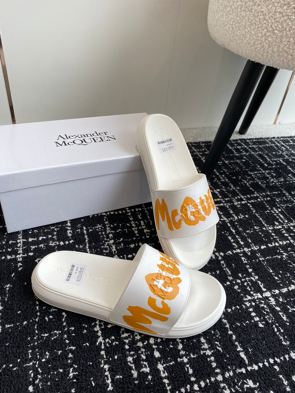 White slide sandals with orange logo strap and padded top, angled on a box.