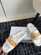 White slide sandals with orange logo strap, pair arranged on a box.