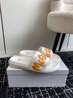 White slide sandals with orange logo strap, shown side by side on a box.