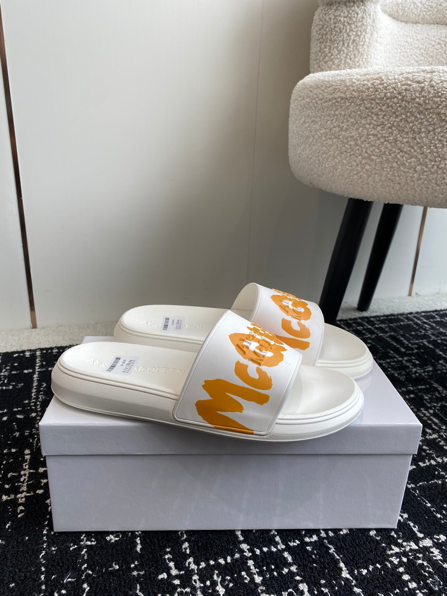 White slide sandals with orange logo strap, shown side by side on a box.