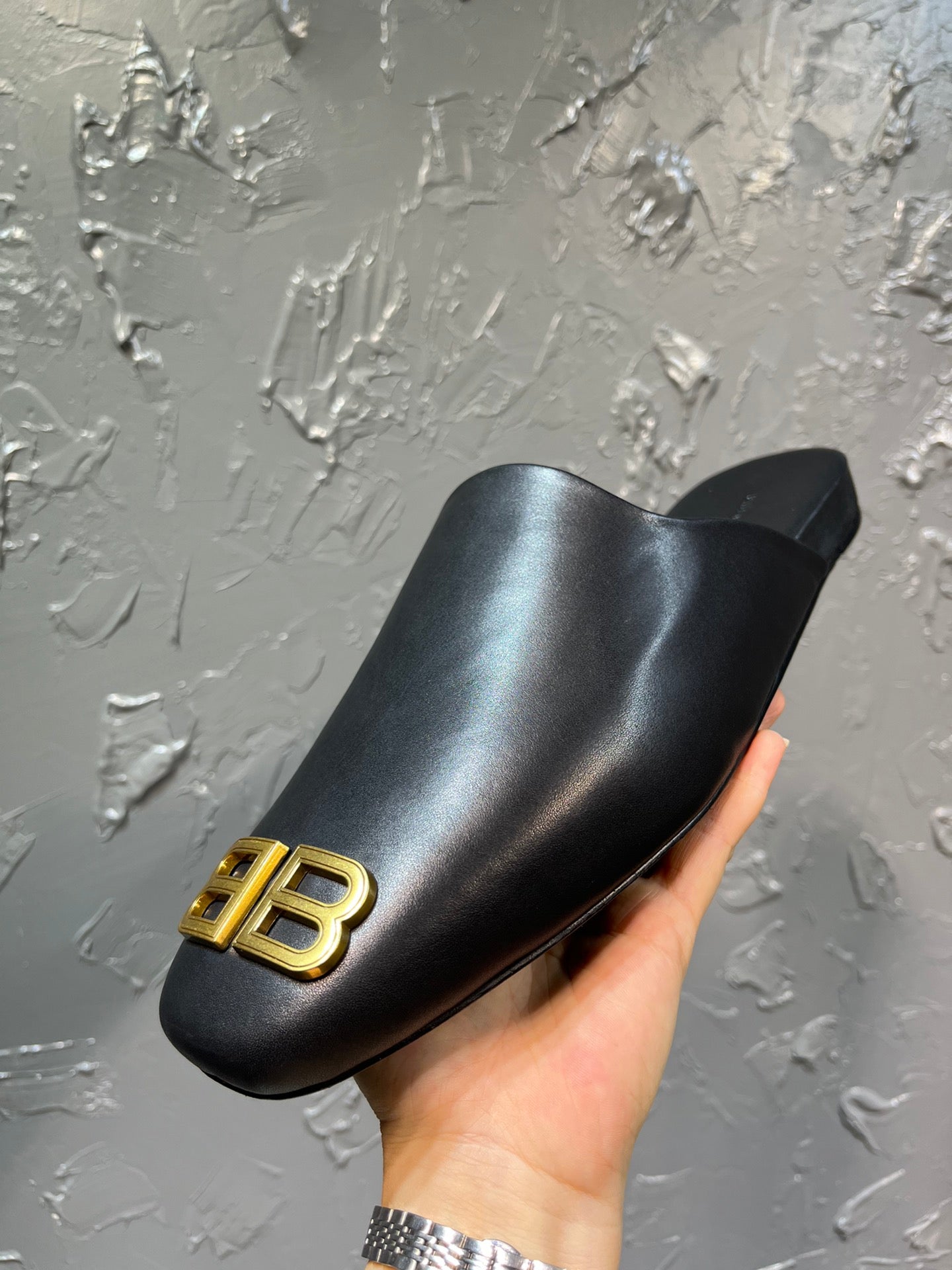 Black slide sandal held in hand with gold BB logo on the front strap.