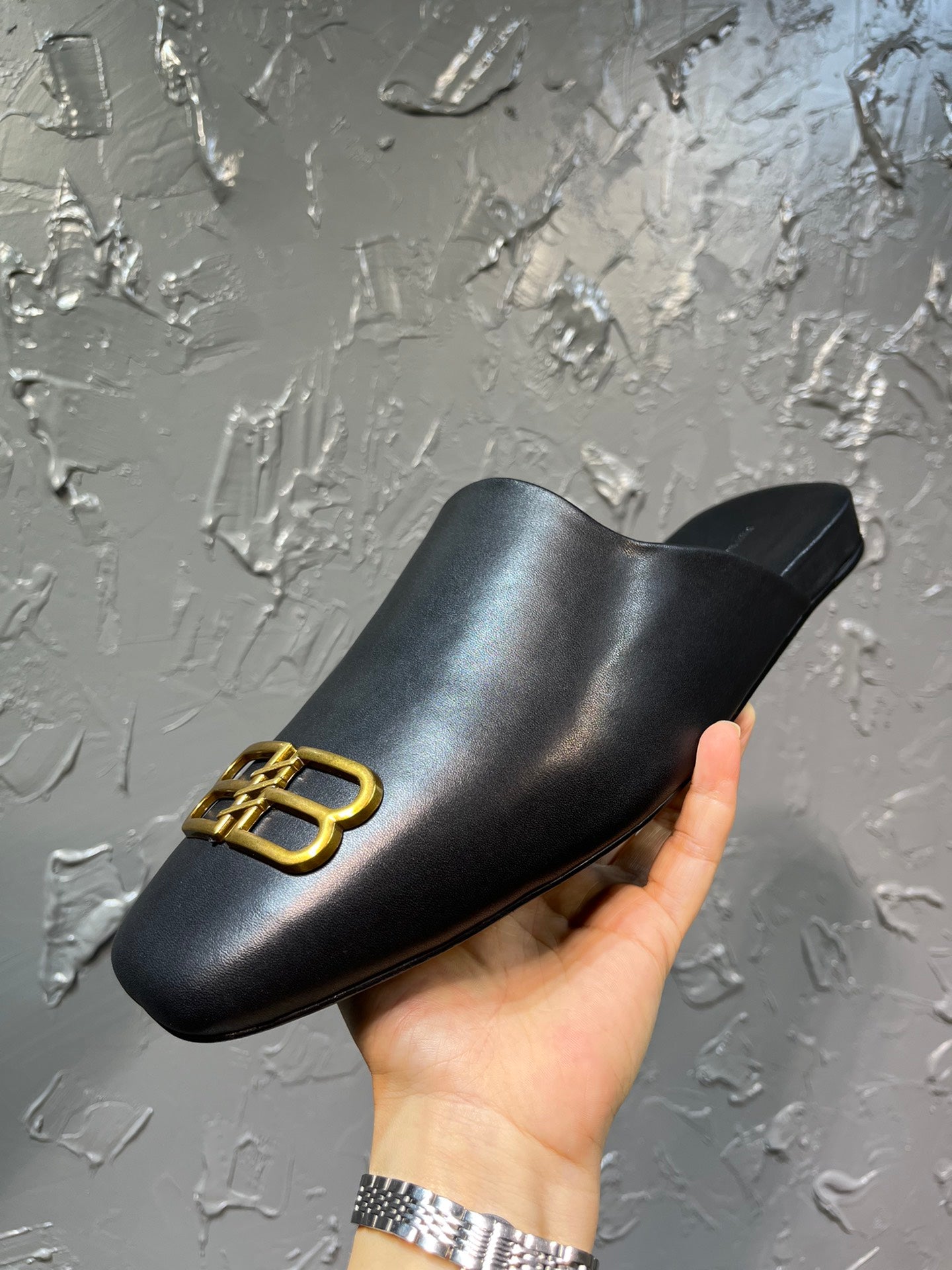 Black leather slide sandal with gold B-logo on upper, side view held up