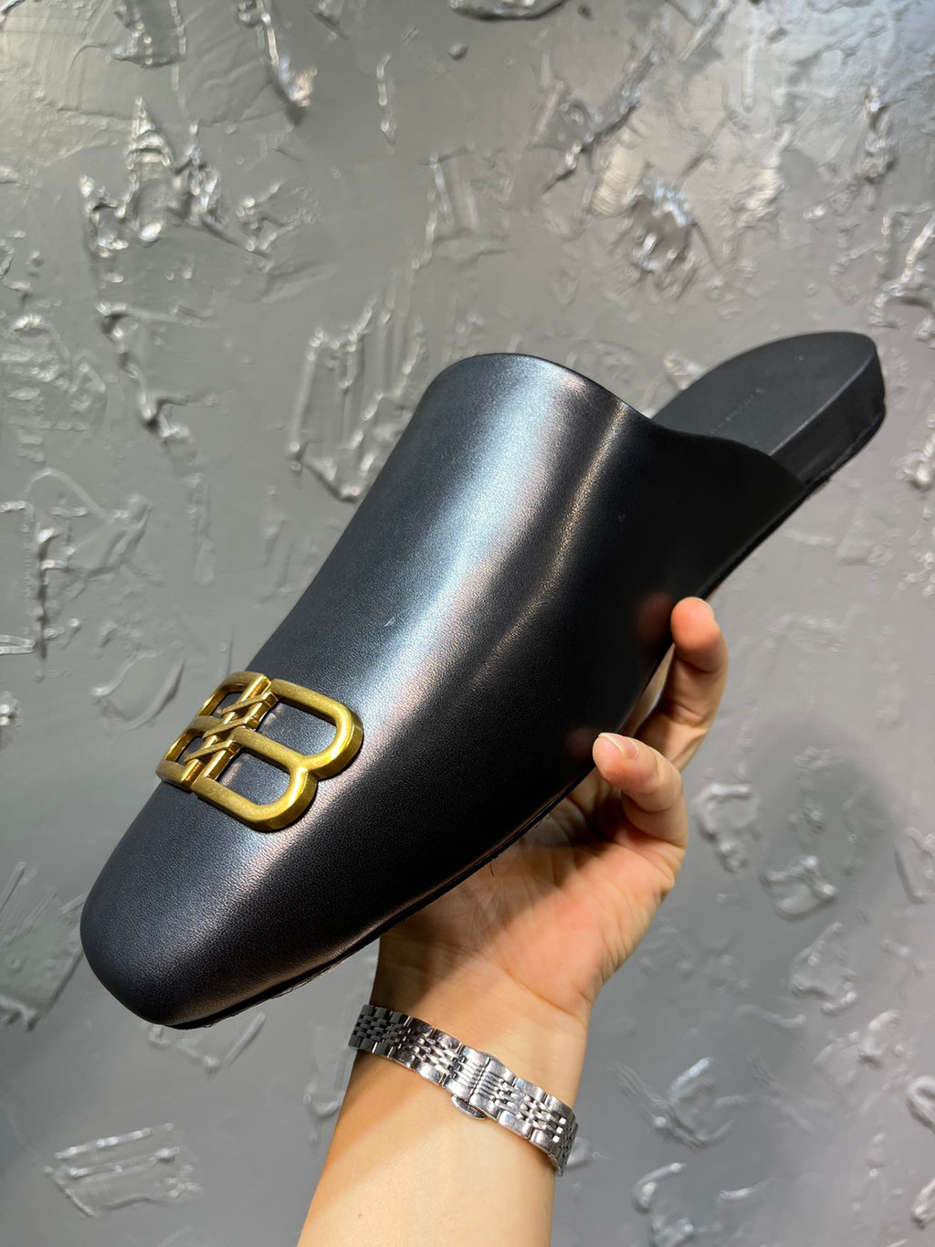 Black leather slide sandal with gold B-logo on upper, held in hand
