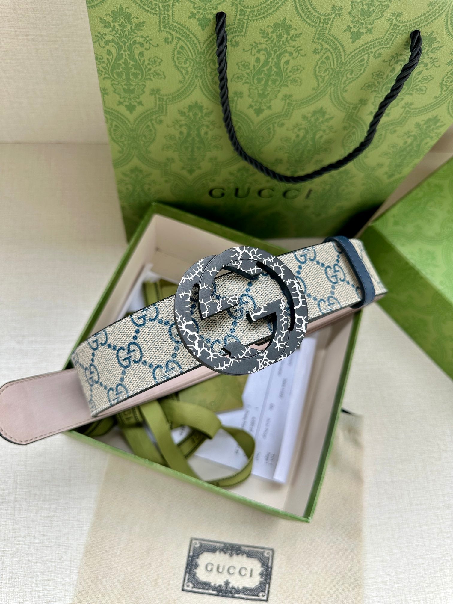 Gucci belt with GG canvas strap and round logo buckle, laid across green gift box.