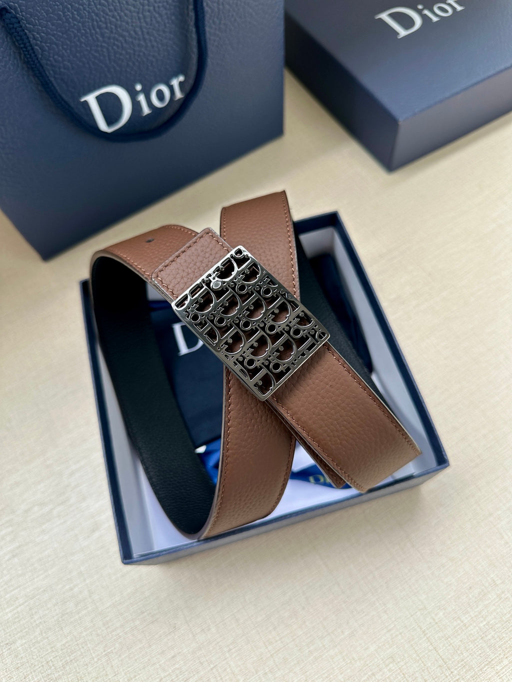 Reversible leather belt in box: brown strap and silver-toned Dior-pattern buckle on top.