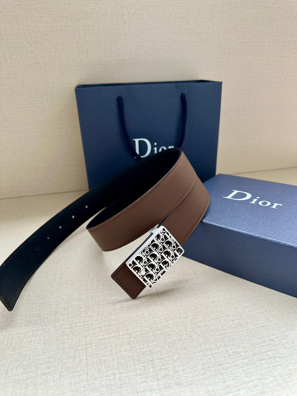 Reversible leather belt with brown strap and silver-toned Dior-pattern buckle, slanted view.