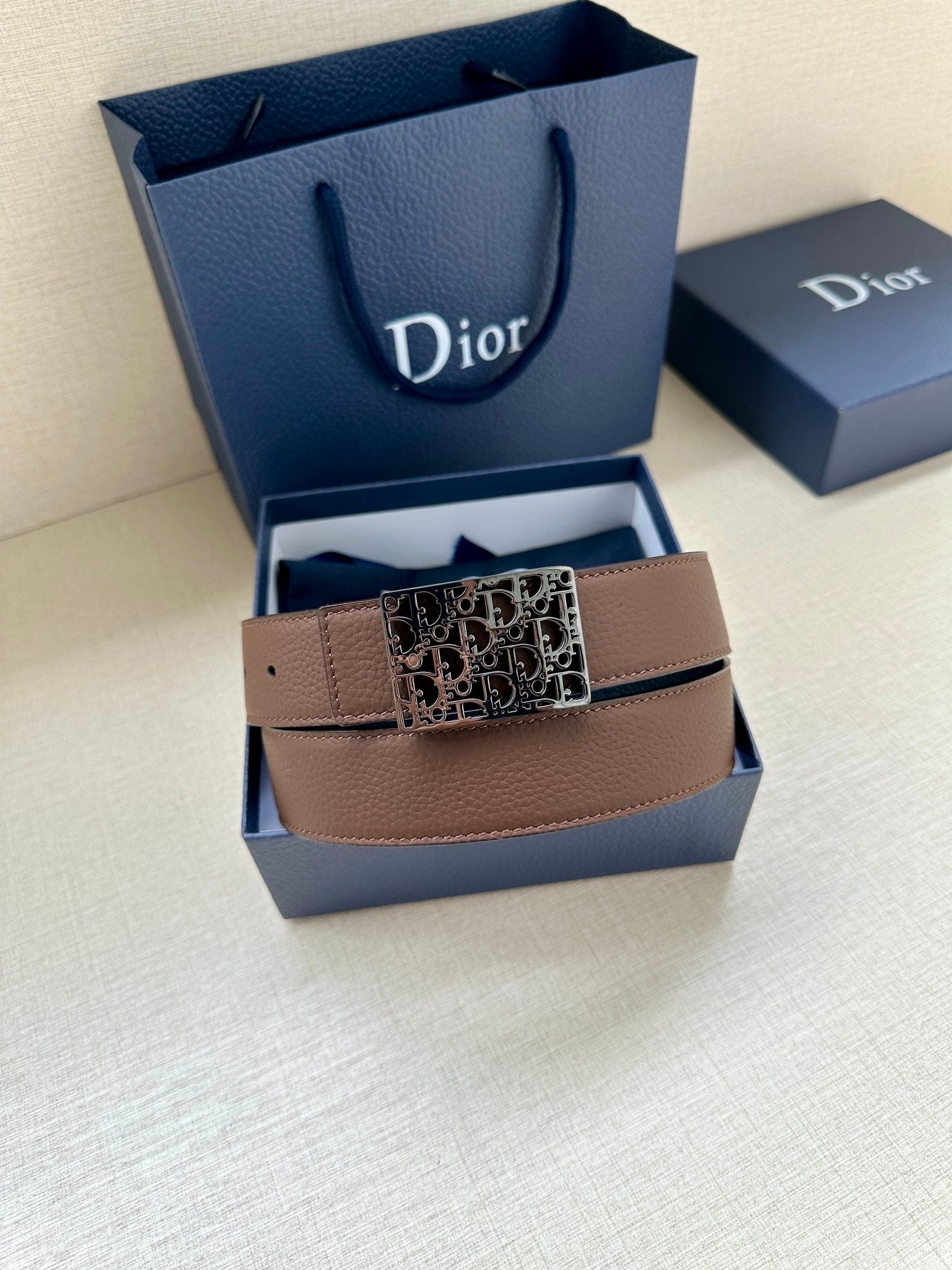 Reversible leather belt with tan/brown strap and silver-toned Dior-pattern buckle, laid in box.