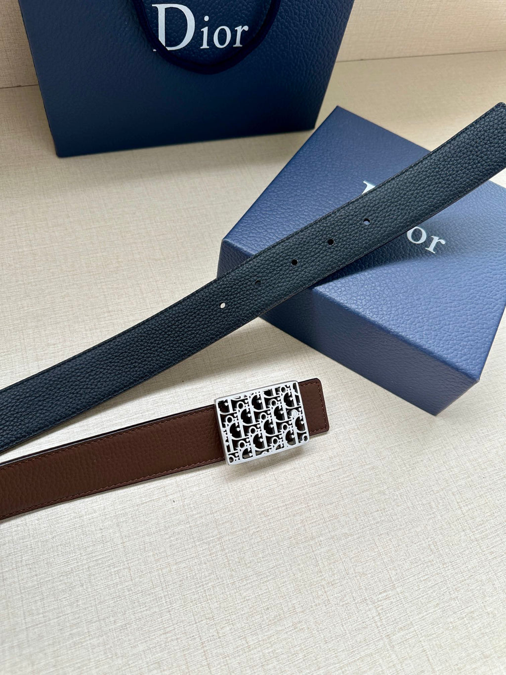 Reversible leather belt with dark strap and silver-toned Dior-pattern buckle, laid across boxes.