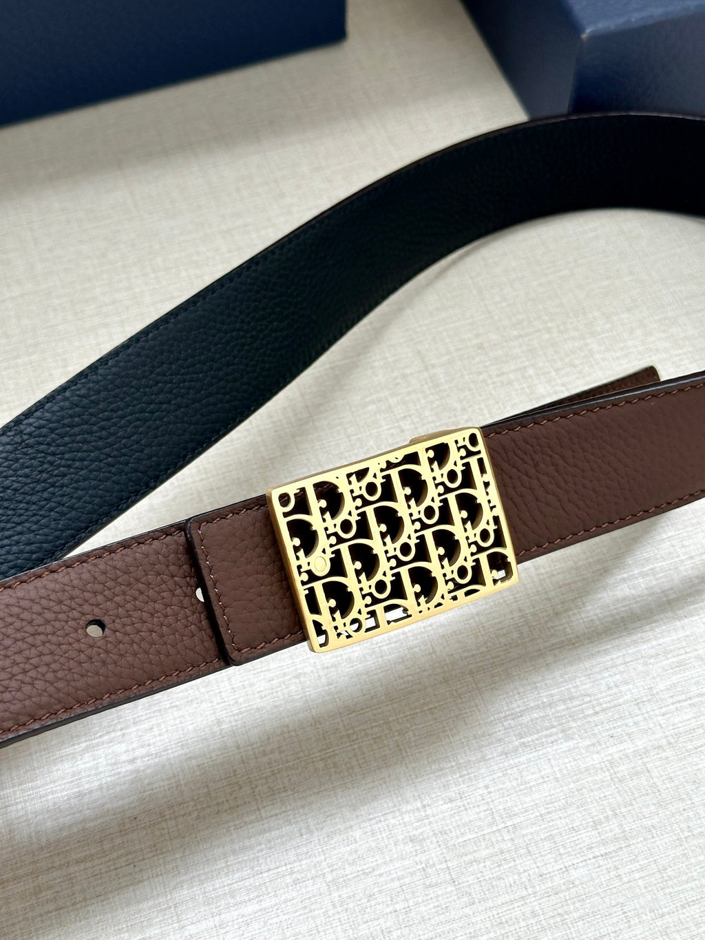 Reversible leather belt close-up: textured black strap, gold Dior-pattern buckle, brown underside.