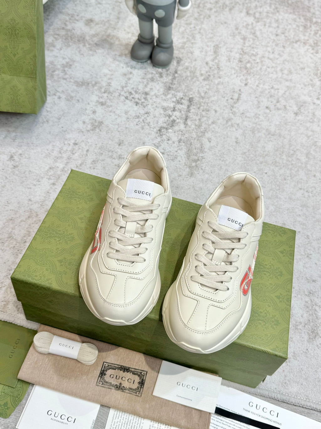 Cream Gucci unisex lace-up sneakers with red GUCCI logo, shown pair on box.