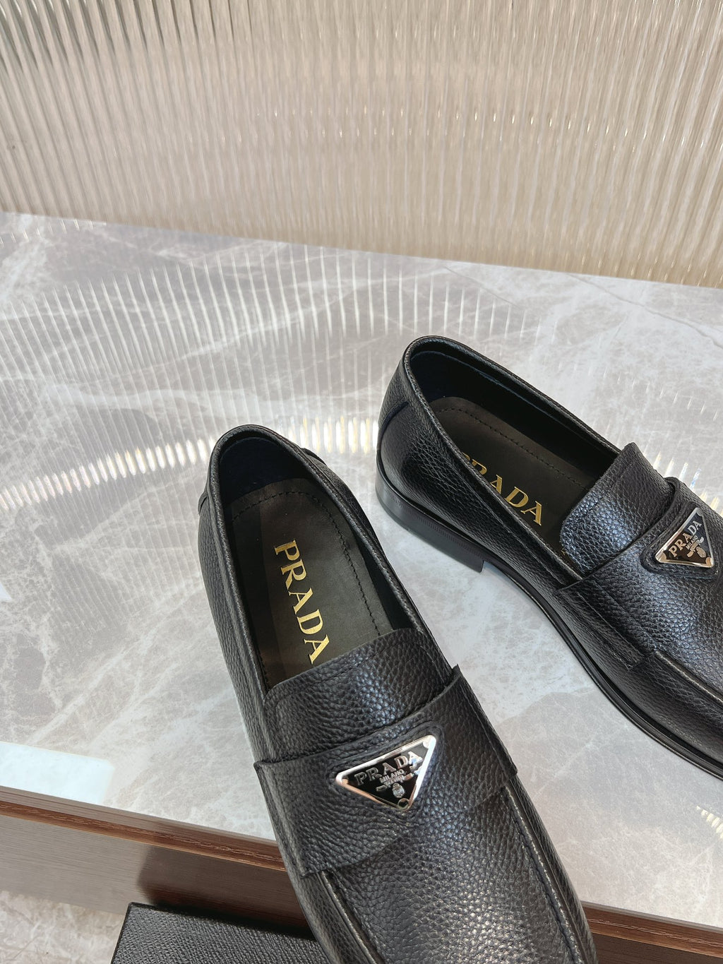 Prada unisex loafers in dark leather, two pairs side-by-side with metal logo badge