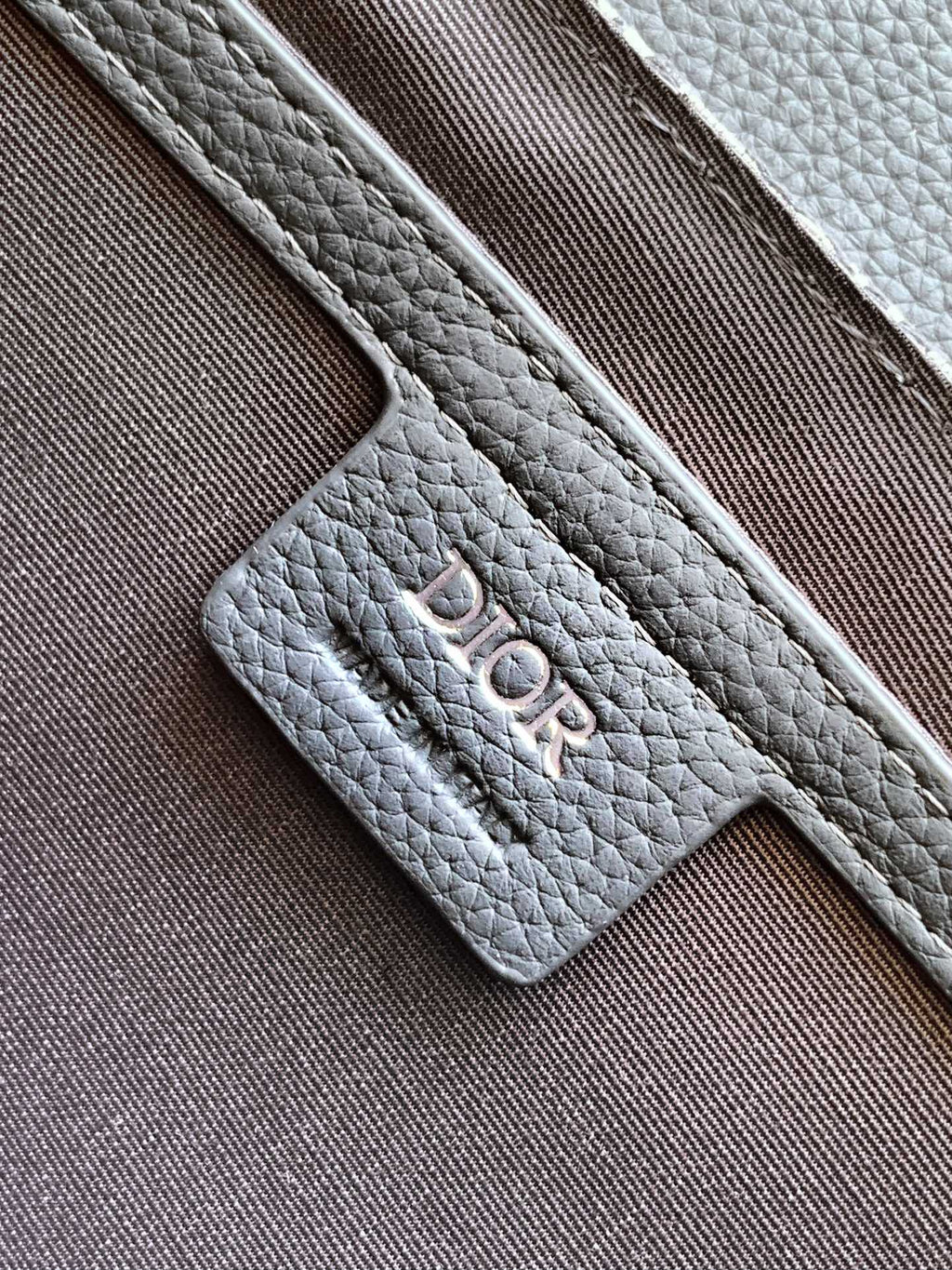 Dior bag strap attachment with embossed logo on gray textured leather.