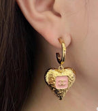 Gold-tone heart drop earring with pink enamel logo on a hoop earlobe.