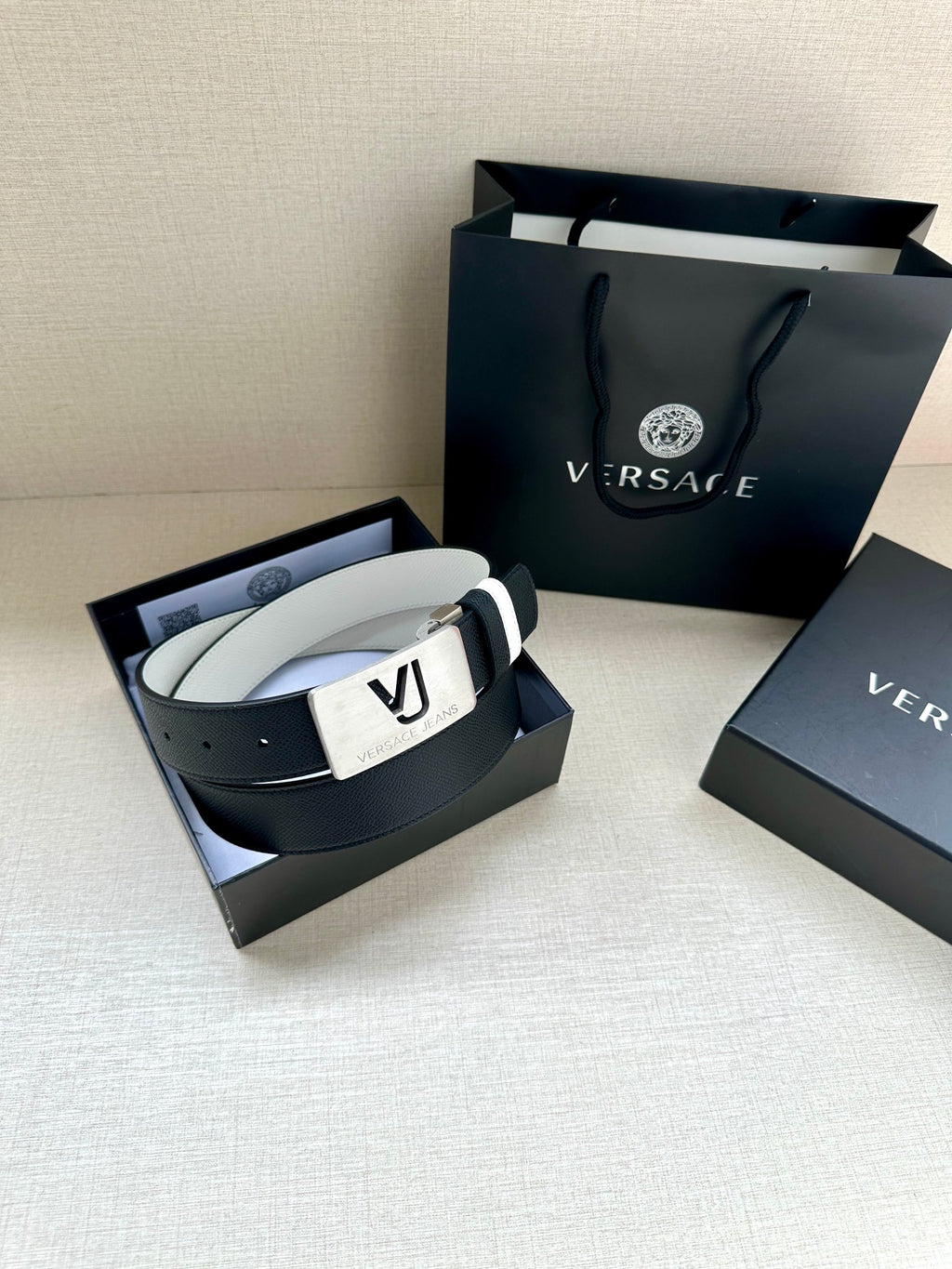 Versace belt in open box: dark strap, white edge stripes, gold VJ logo buckle