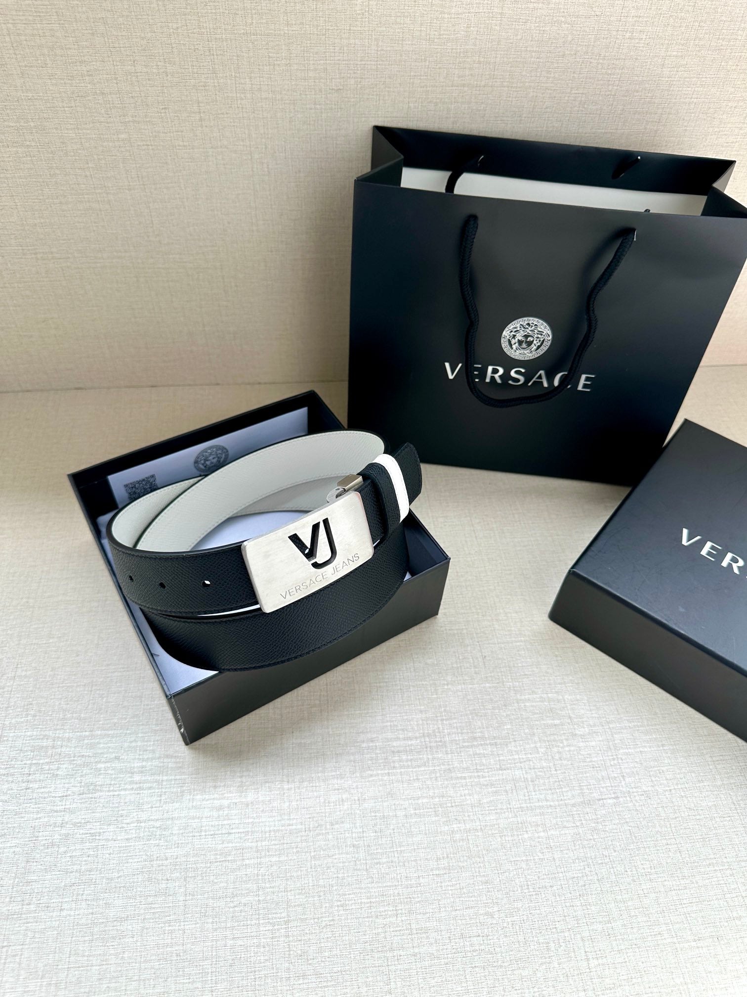 Versace belt in open box: dark strap, white edge stripes, gold VJ logo buckle