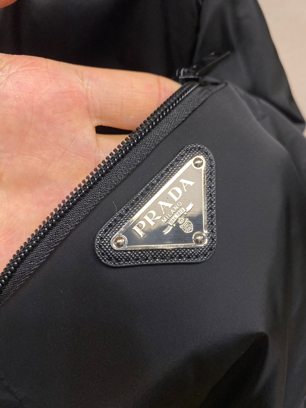 Prada long nylon jacket, dark color, close-up of triangular logo patch on pocket