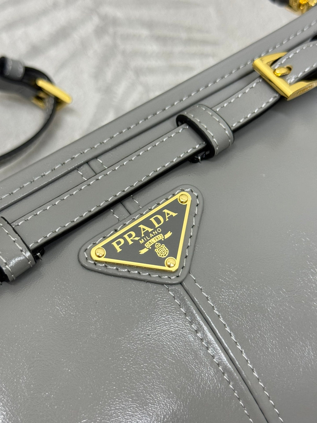 Prada bag strap close-up with gold buckle and triangular logo patch on gray leather.