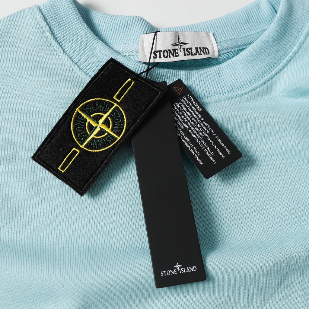 Light blue crewneck sweatshirt with black front logo patch and attached tags