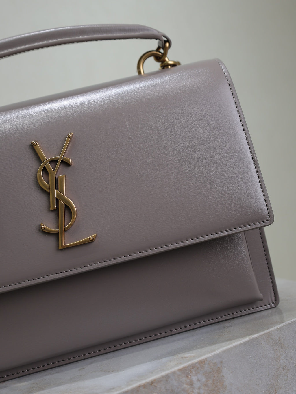 YSL bag close-up showing front flap edge, gold-tone YSL logo, and top handle, gray-lavender
