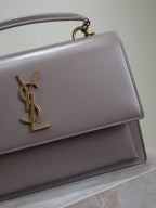 YSL bag close-up showing front flap edge, gold-tone YSL logo, and top handle, gray-lavender
