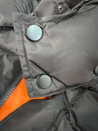 Long puffer jacket front with round snap buttons and orange lining edge.