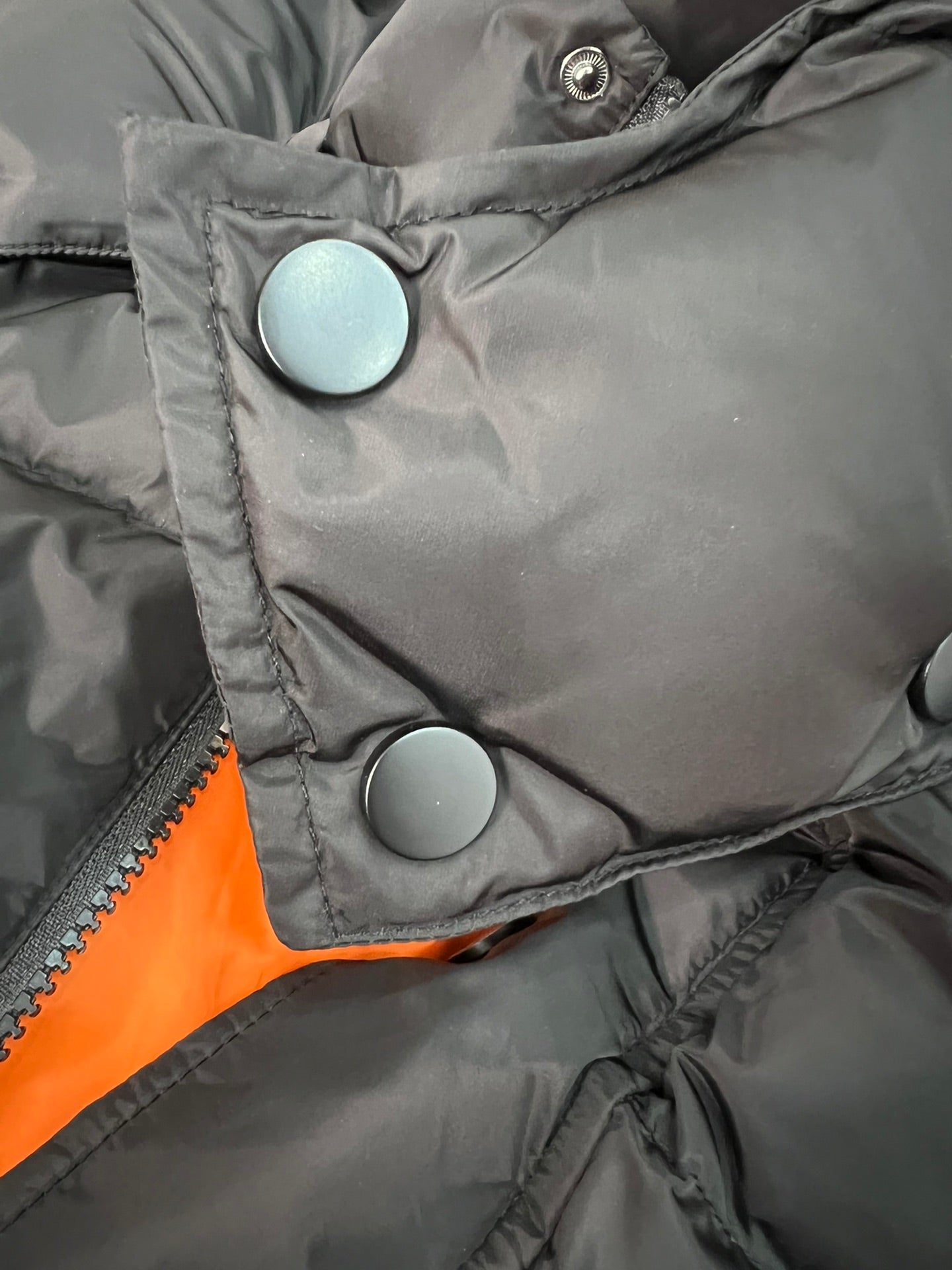 Long puffer jacket front with round snap buttons and orange lining edge.
