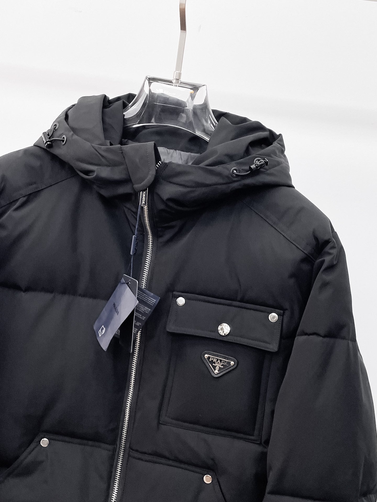 Black hooded puffer jacket front close-up with zipper, logo patch, and flap chest pocket
