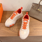 White and orange Hermes-style sneakers with lace-up closure and textured rubber sole.