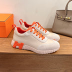 White sneakers with orange laces, side panel stitching, and chunky white rubber sole.