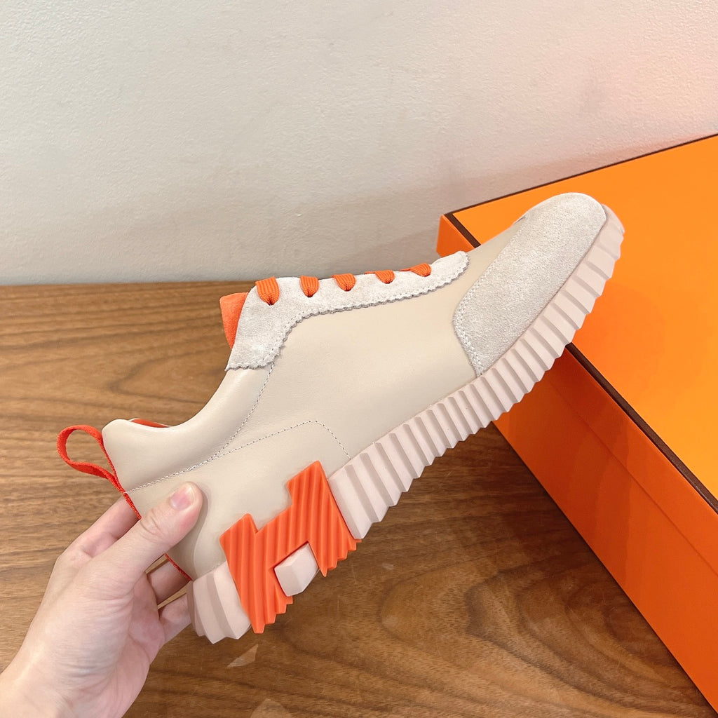Side view of white sneakers with orange-trimmed sole and ribbed traction tread.