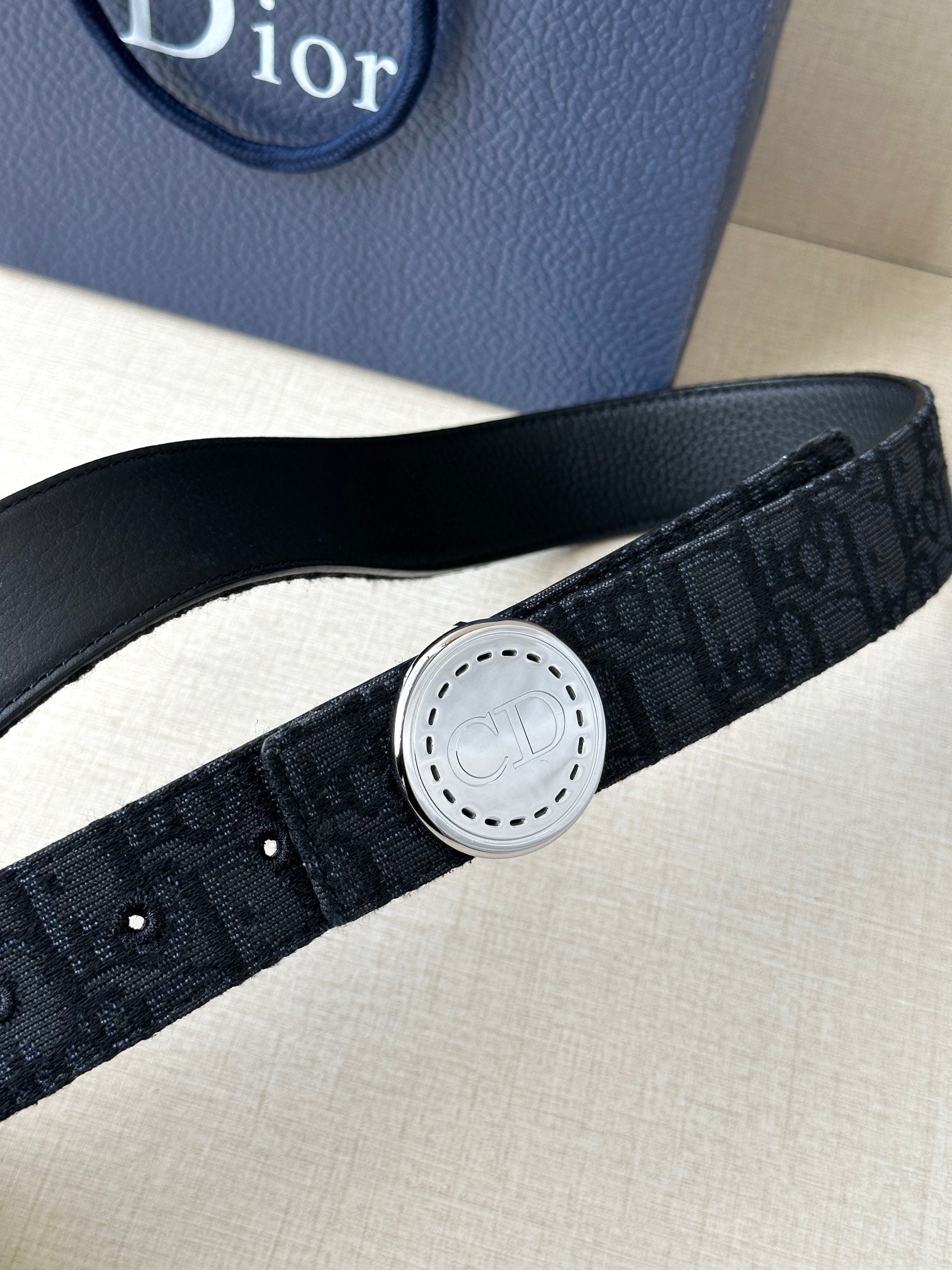 Dior belt close-up showing dark monogram strap and round silver-tone logo buckle