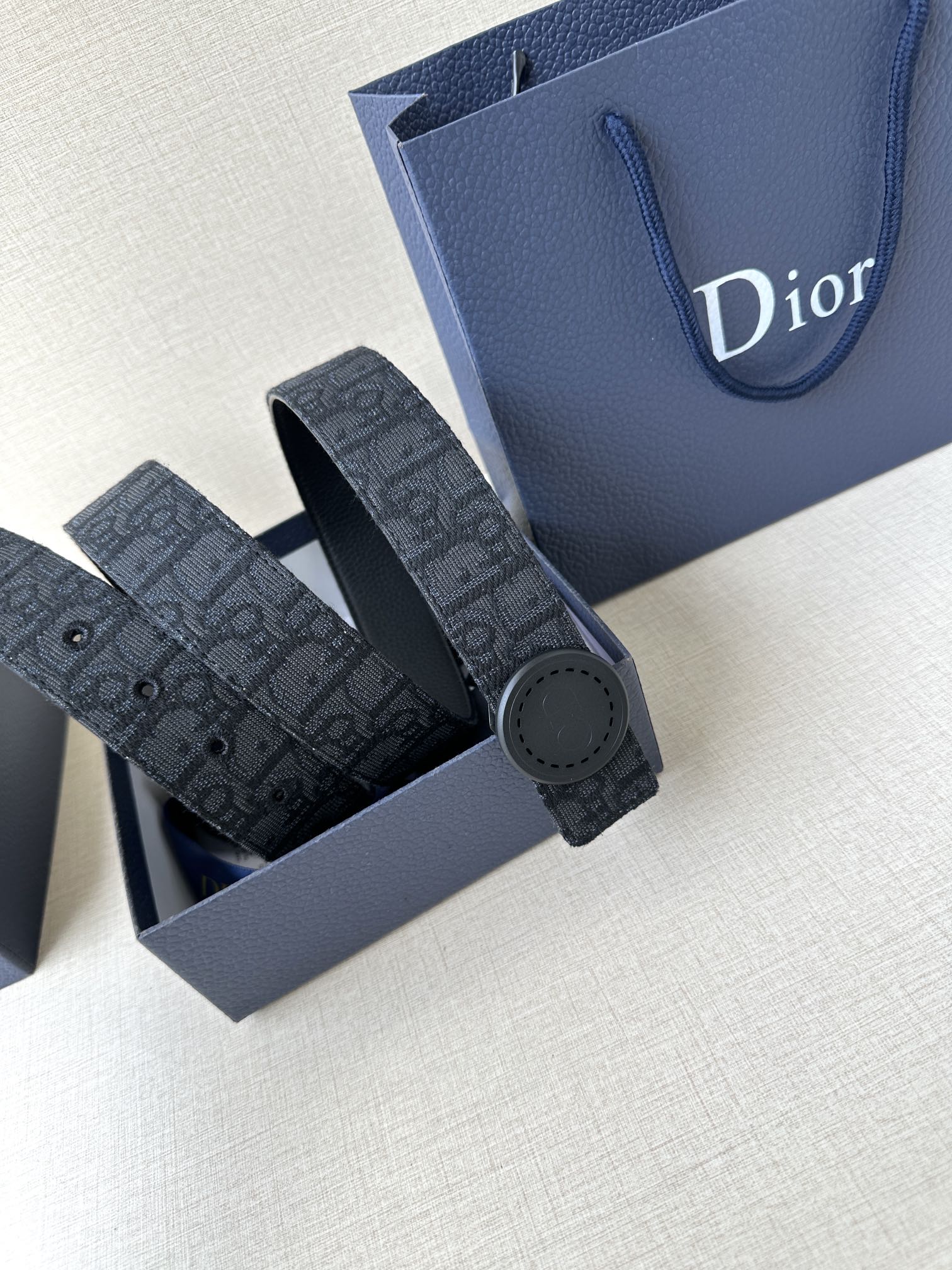Dior belt with dark monogram strap and round gray buckle, stacked on blue box