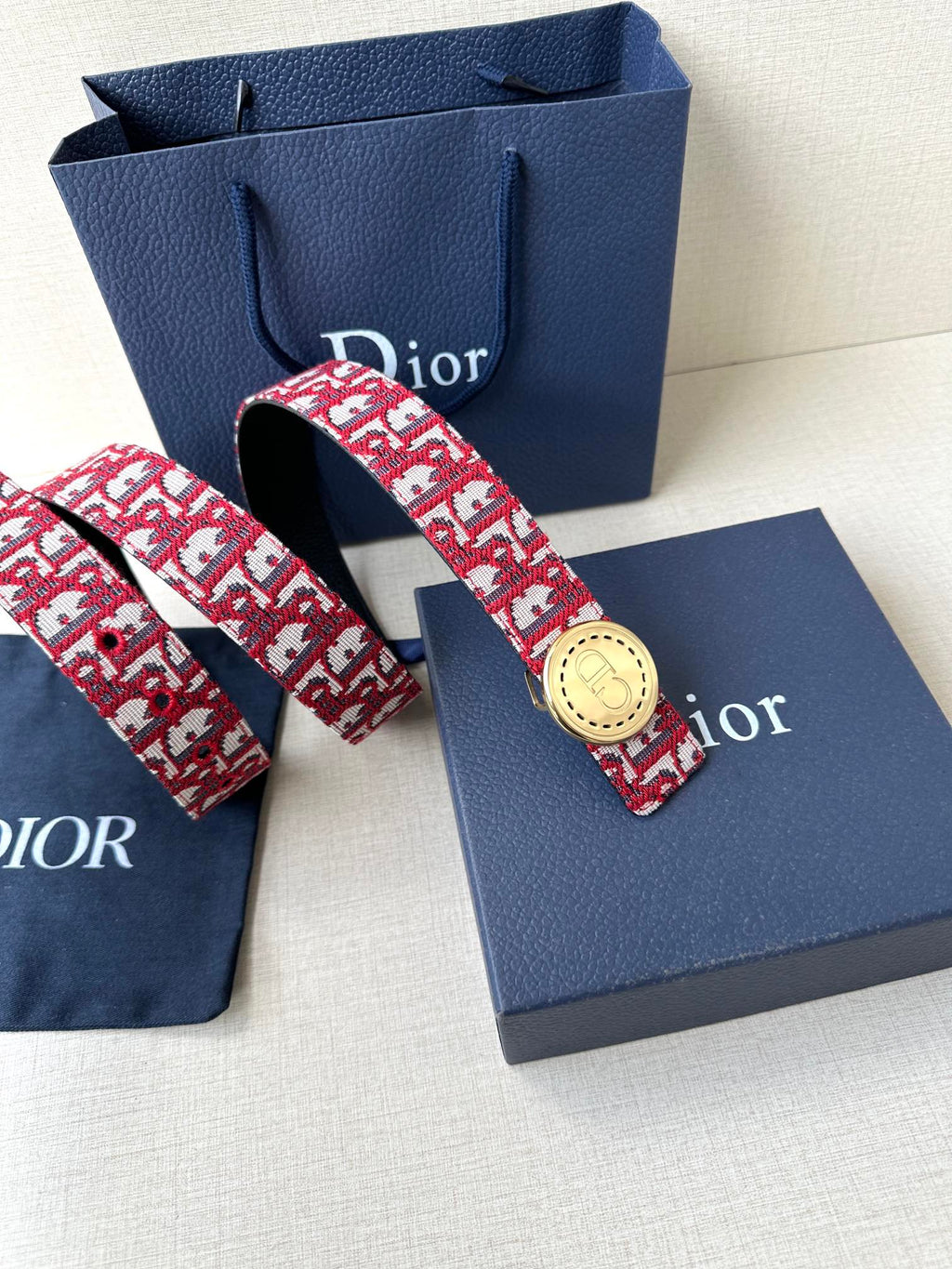 Dior belt displayed with blue box; red-and-white monogram strap and round gold buckle