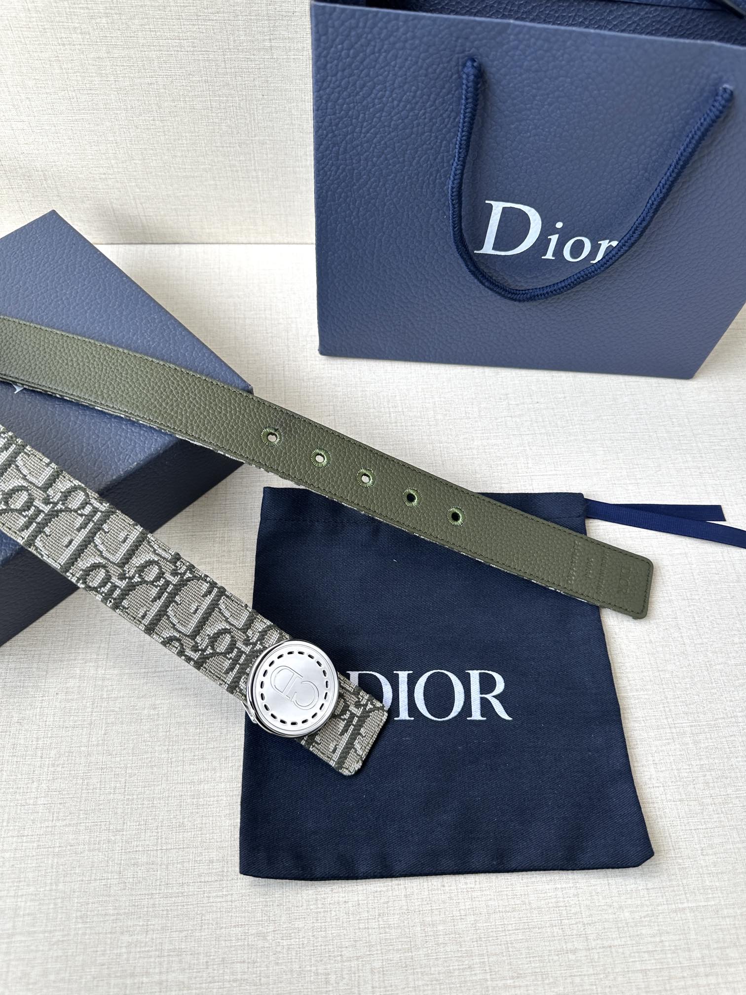 Dior belt with monogram strap and round light buckle, styled on a blue gift bag.