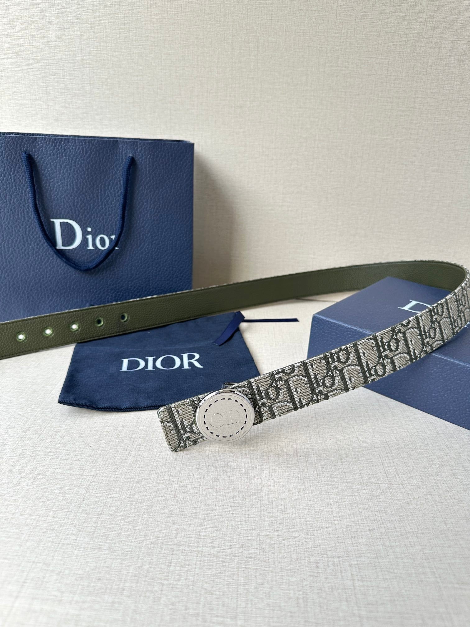 Dior belt with monogram fabric strap and circular pale buckle, laid across a box.