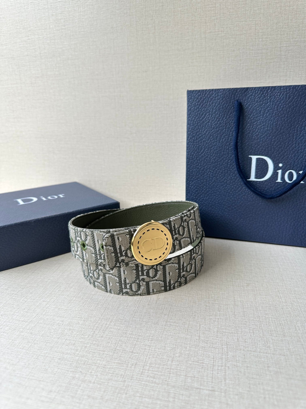 Dior belt with monogram fabric strap and round tan-gold buckle, against a blue box.