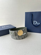 Dior belt with monogram fabric strap and round tan-gold buckle, against a blue box.