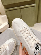 White low-top sneaker with black logo patch and stitched toe, lace-up, angled view.
