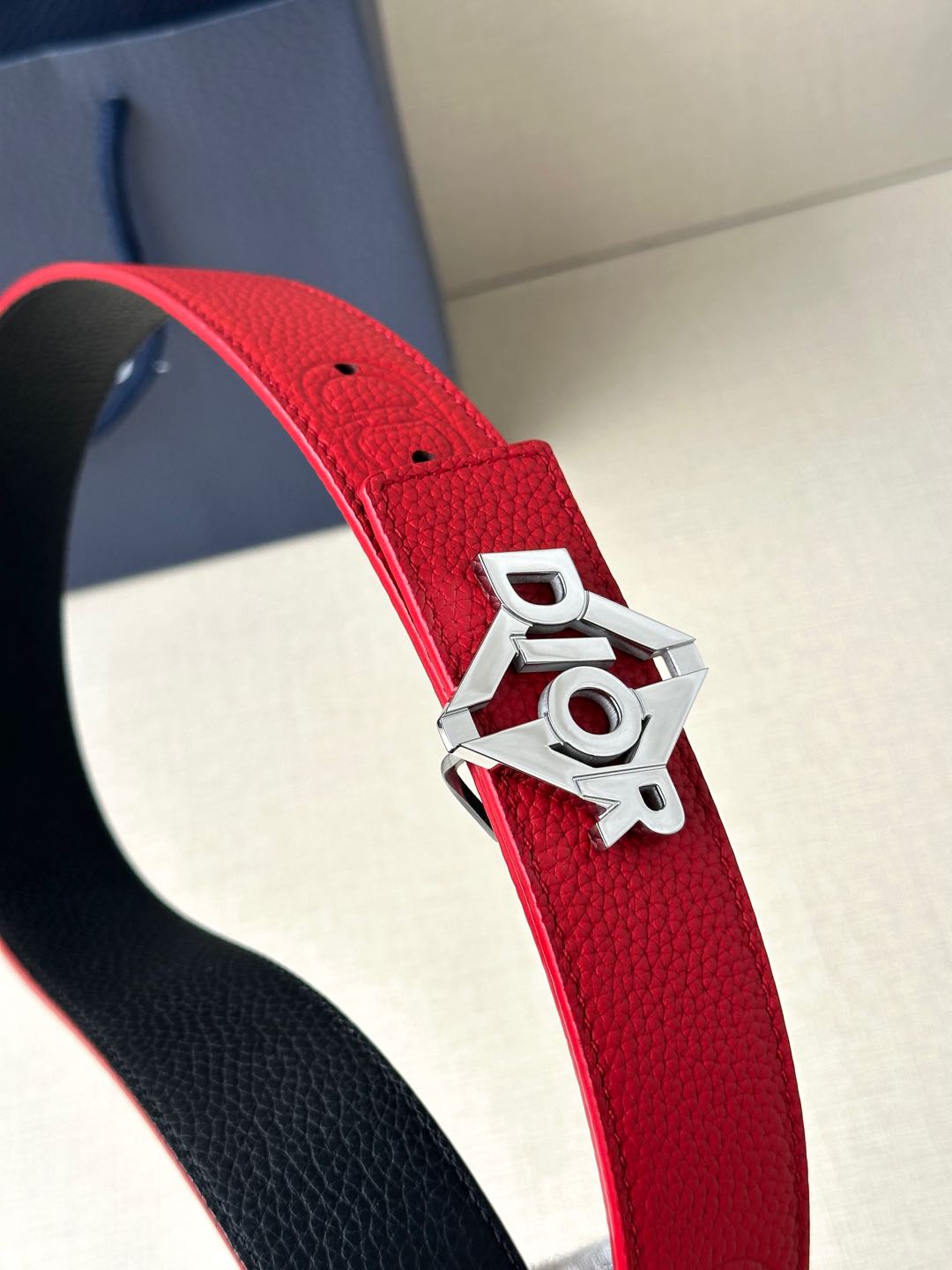 Red leather belt strap with silver buckle featuring Dior lettering and adjustable holes.