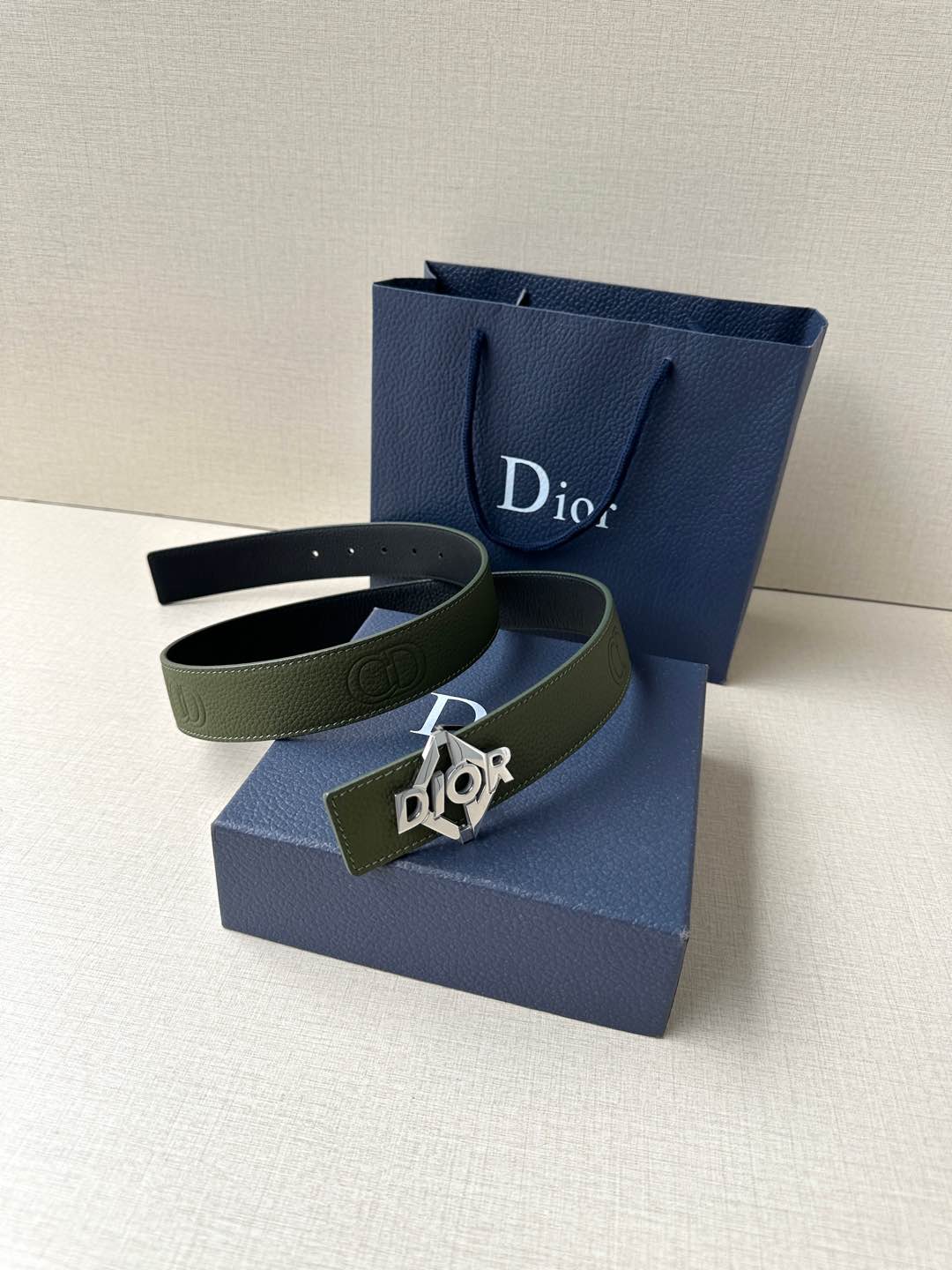 Dior leather belt shown with olive strap and dark logo buckle on a bag stand