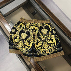 Black and gold baroque patterned shorts with Greek key border, laid flat in a box.