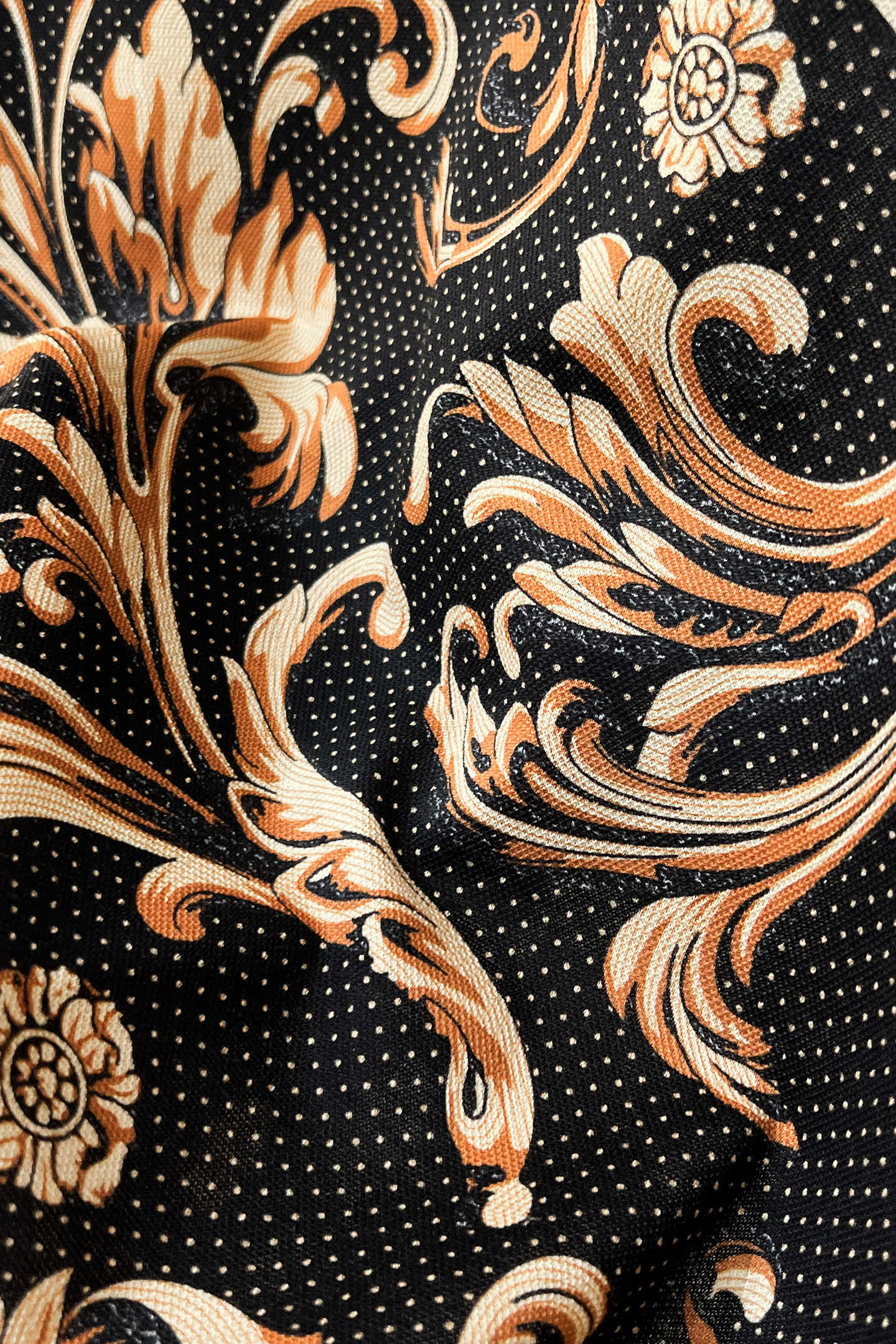 Shirt fabric close-up: black ground with gold-and-orange floral scrolls and tiny dot pattern.