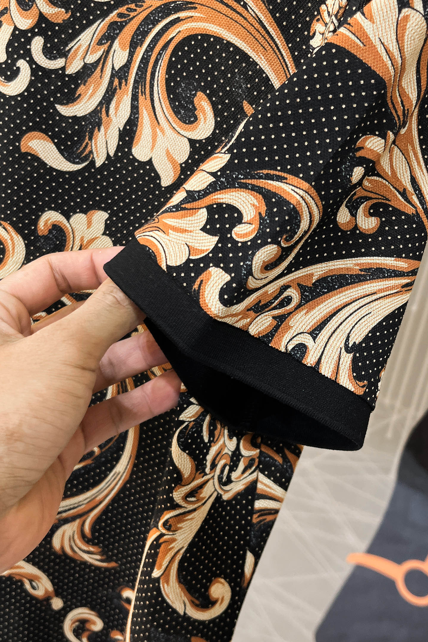 Shirt cuff close-up: black sleeve edge with baroque print in gold and orange on knit fabric.