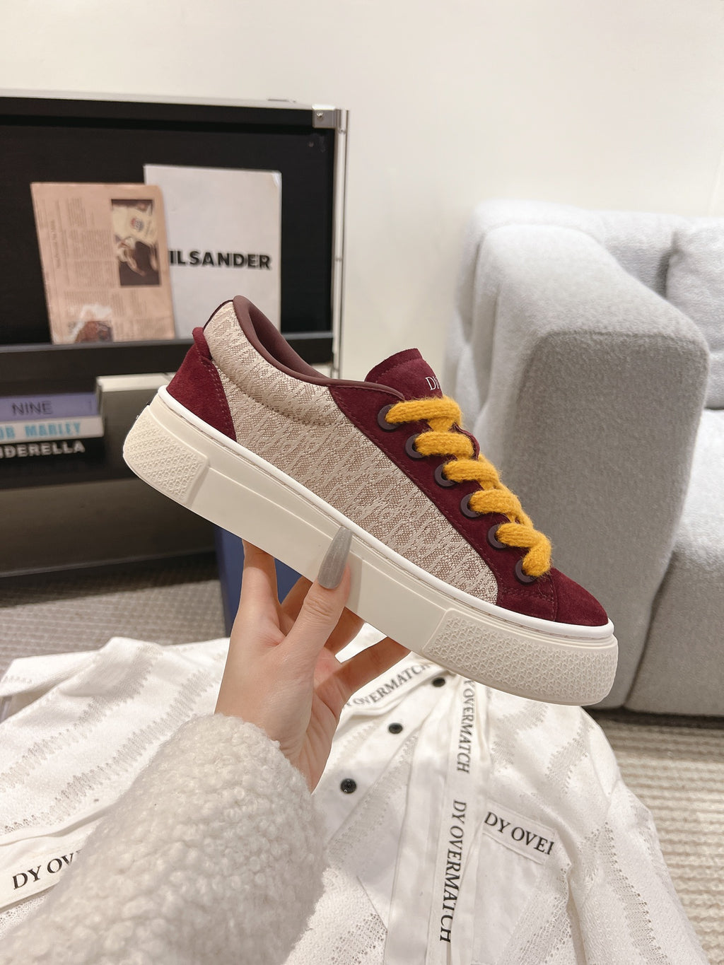 Burgundy canvas-style sneaker with yellow laces and white sole, side view held by hand.