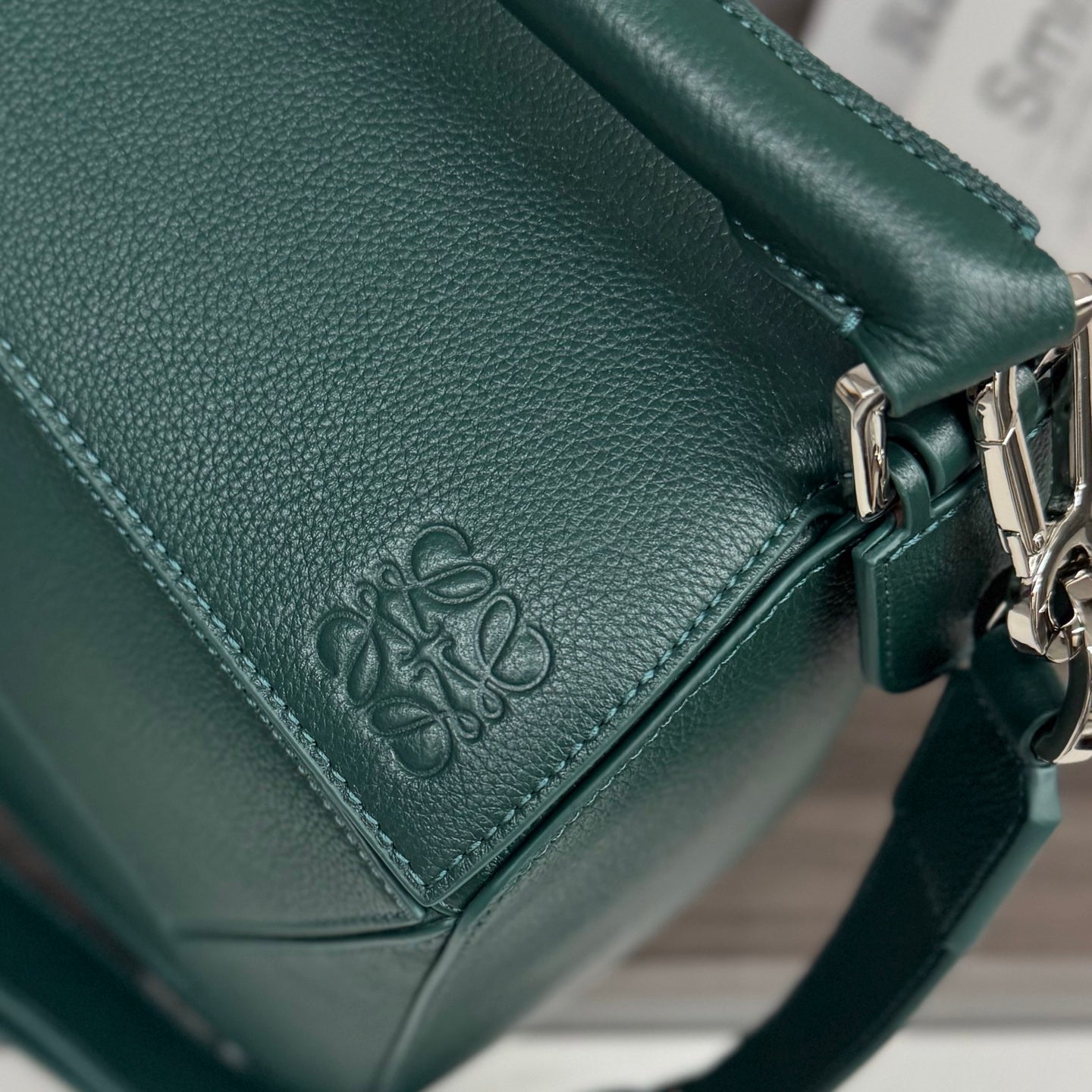 Teal leather bag corner with embossed logo, stitched seam detail, and metal ring hardware.