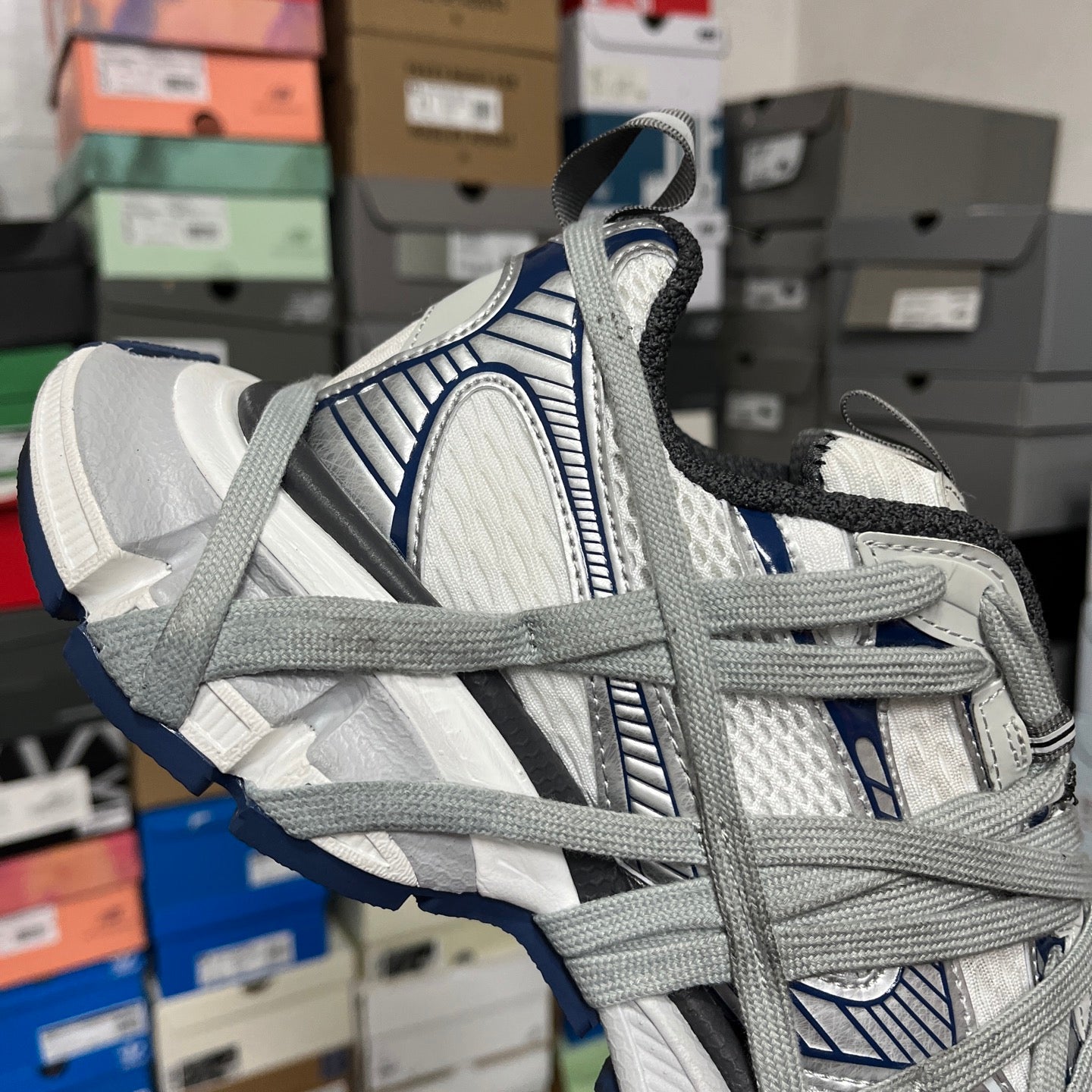 Side view of grey and white athletic shoe with blue logo detail and lace overlay.